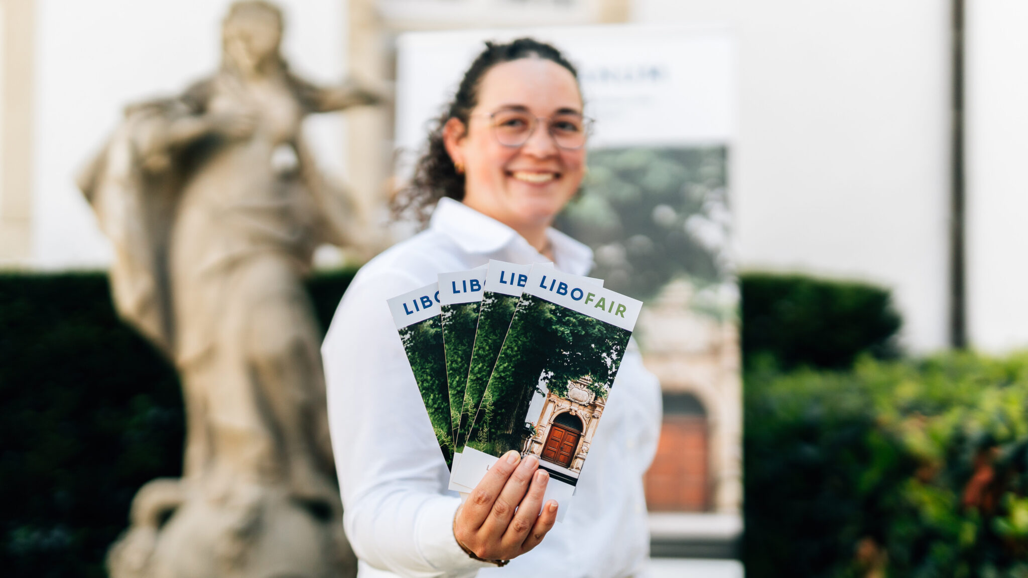 Eine Dame in weißer Bluse hält die Flyer für die Libofair in der Hand. Im Hintergrund ist verschwommen ein Roll up und eine Statue erkennbar.